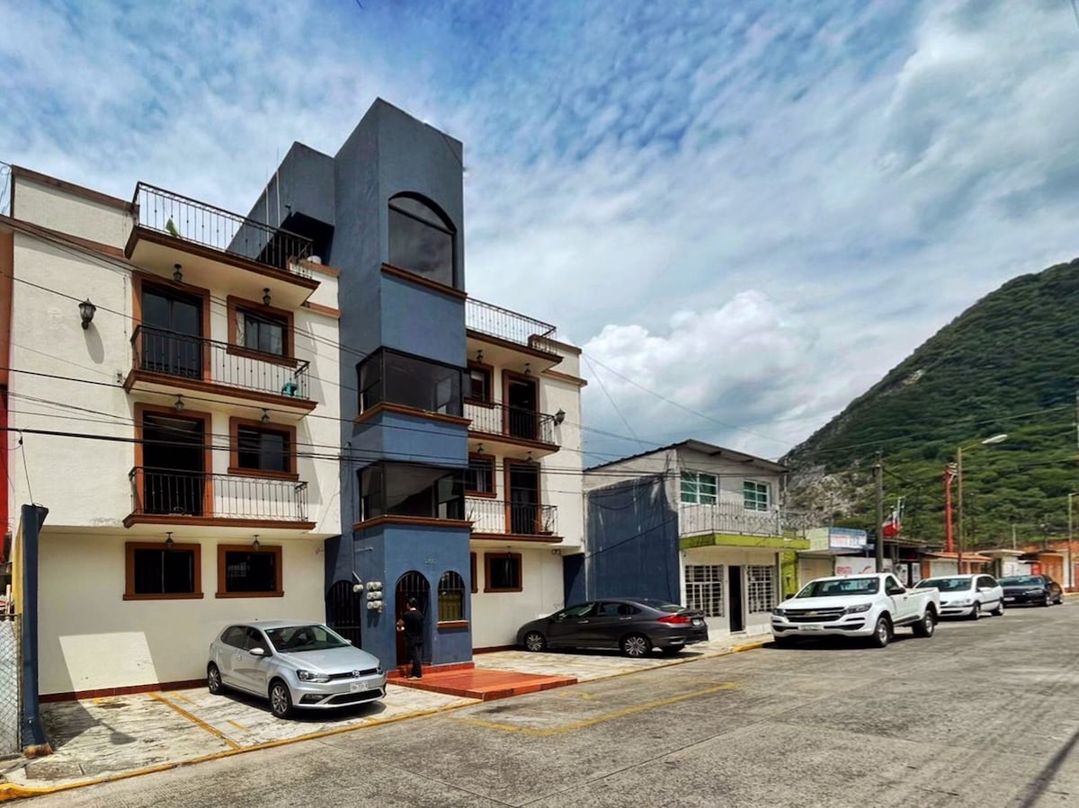 A three-story building with a modern design is seen on a quiet street. The structure features a combination of dark and light exterior colors, with balconies on the upper floors. Parking spaces for several cars are visible in front.