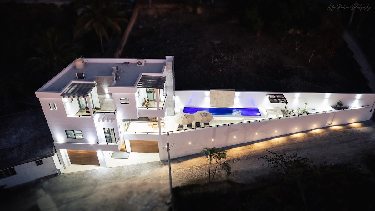 An aerial view of a modern, spacious house is presented, featuring a large pool surrounded by outdoor seating. The exterior is illuminated, emphasizing clean lines and a stylish design. The property is flanked by a driveway and lush greenery, conveying a tranquil environment.