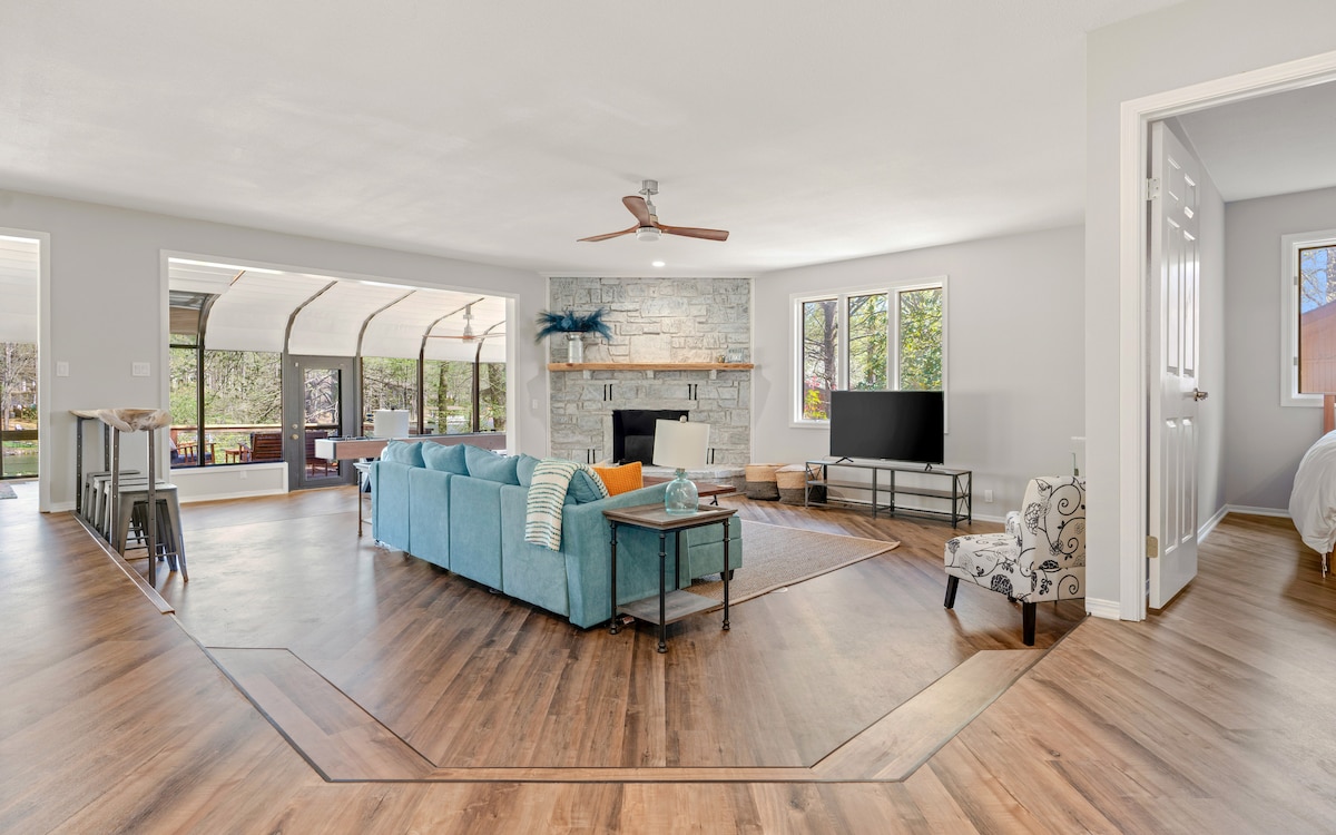 The open-plan living area features a spacious sectional sofa in a calming blue hue, complemented by warm wood flooring. A modern stone fireplace serves as a focal point, and large windows provide natural light and views of the outdoors. A smart TV is positioned for entertainment.