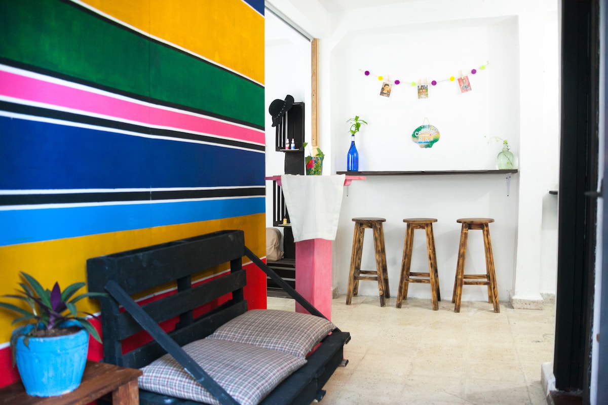 A vibrant wall with horizontal stripes in various bold colors creates an inviting atmosphere. A simple bar area is equipped with three wooden stools, while a potted plant adds a touch of greenery. Warm light enhances the open space leading to other areas of the accommodation.