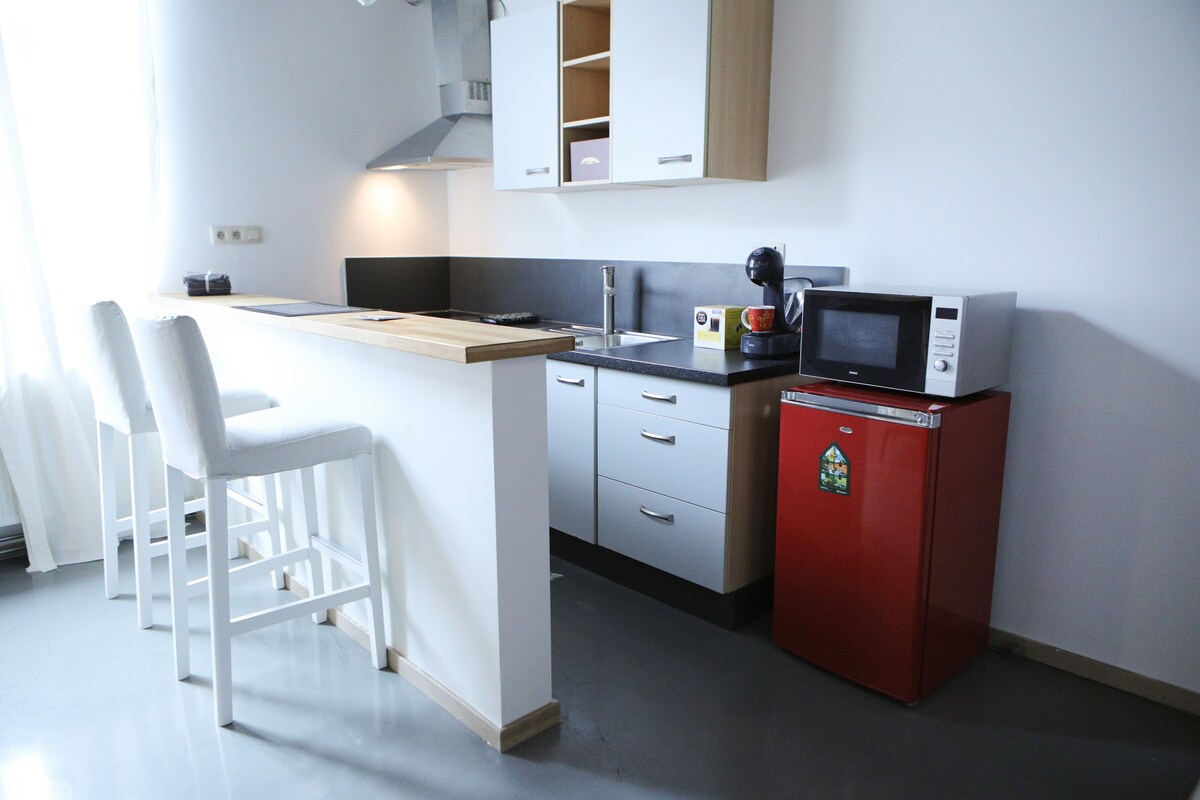 A modern kitchen features a wooden countertop with two high stools positioned at one side. A sleek range hood is mounted above the stove. Below the counter, a red refrigerator and various kitchen appliances are visible, complemented by a clean and minimalist aesthetic.