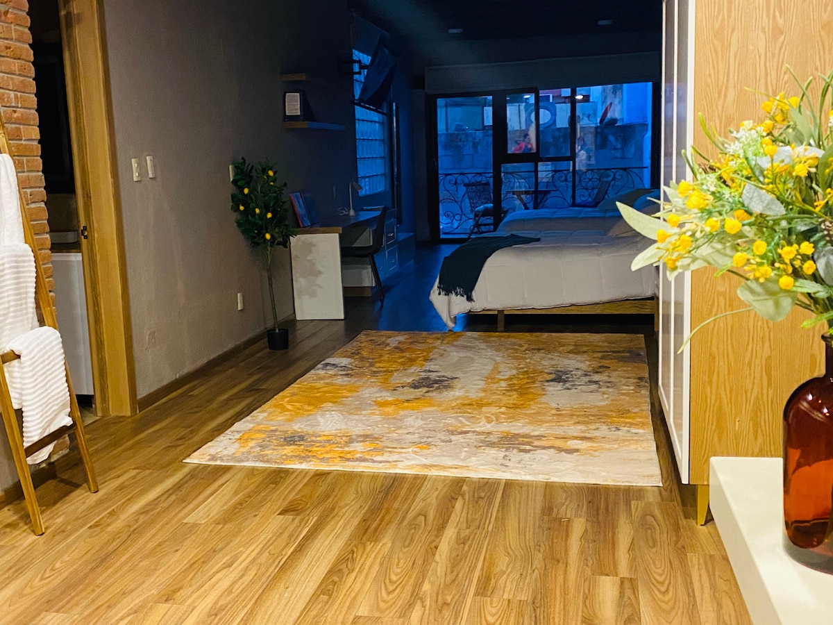 A cozy interior space features a light-colored area rug on warm wooden flooring. In the background, a bright bedroom with a spacious bed is visible, complemented by natural light from large windows. A decorative arrangement of flowers is situated on a nearby surface.