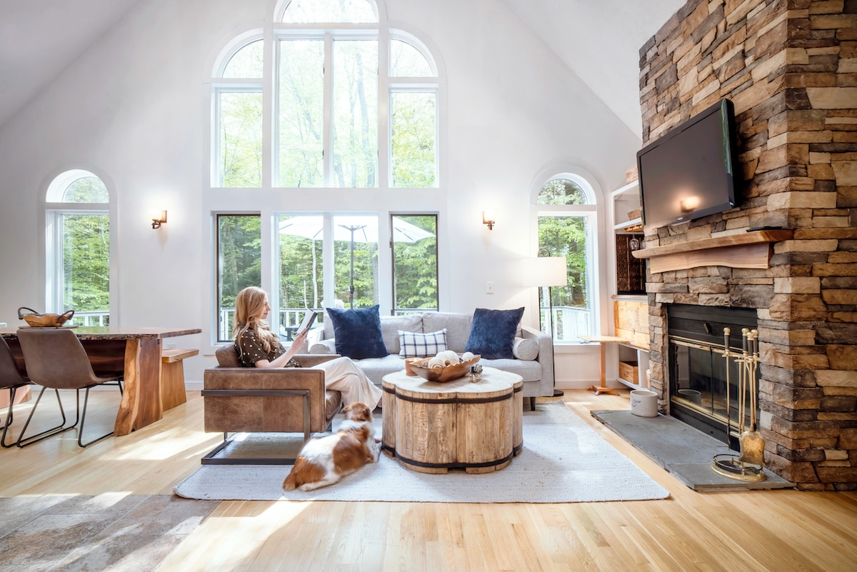 A spacious living area features large windows that allow natural light to fill the room, highlighting a stone fireplace and wooden accents. A comfortable sofa is arranged near a coffee table, with a dining area visible in the background, creating a cozy gathering space.