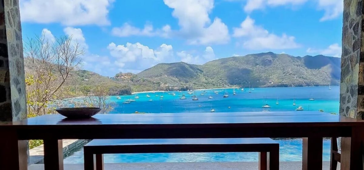 A scenic outdoor view features a spacious wooden table overlooking a stunning seascape. The vibrant blue waters of the harbor are dotted with sailboats, framed by lush green hills and a partly cloudy sky.