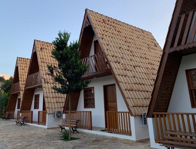 A row of charming chalets features distinct A-frame roofs and wooden details, set against a calm background. Each chalet is positioned with a small porch and outdoor seating, providing a cozy entryway. Lush greenery is visible, enhancing the overall natural setting.