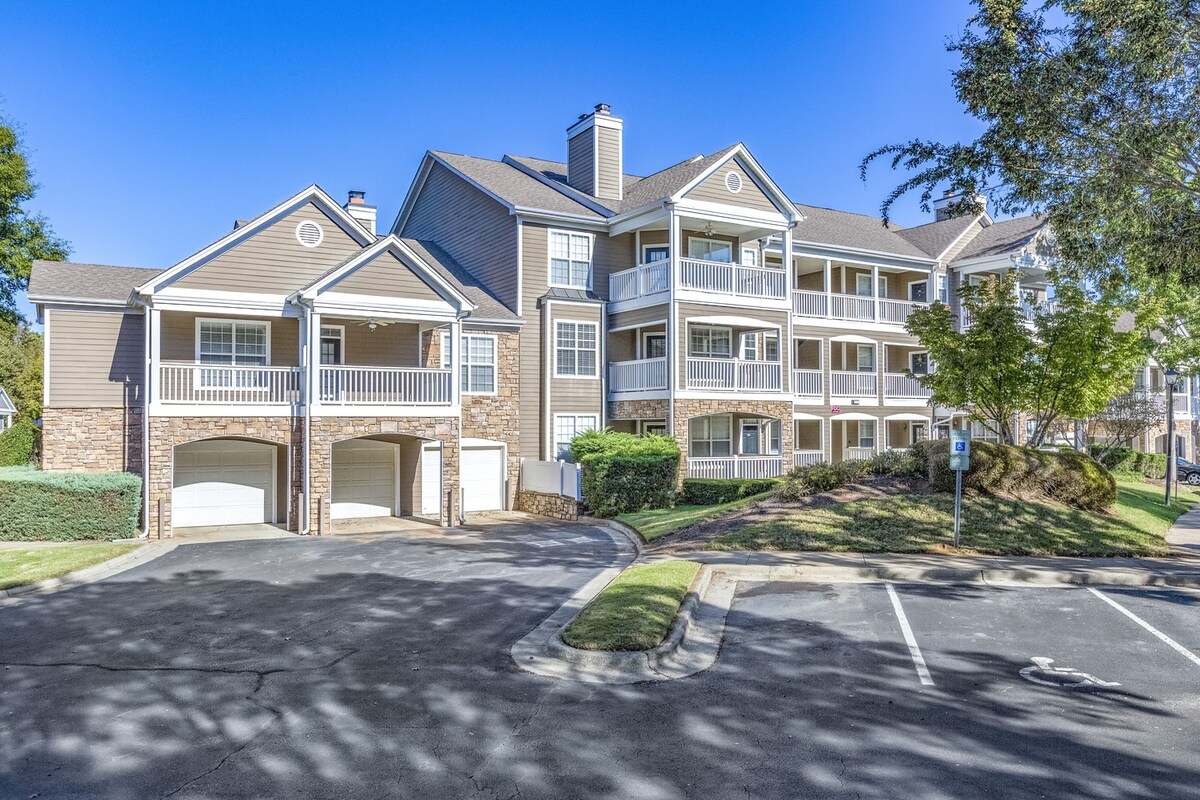 The exterior view features a multi-story apartment building with a combination of stone and wood siding. Balconies are visible on the upper levels, framed by trees and manicured landscaping. A driveway and parking spaces are situated in the front, contributing to the inviting community atmosphere.