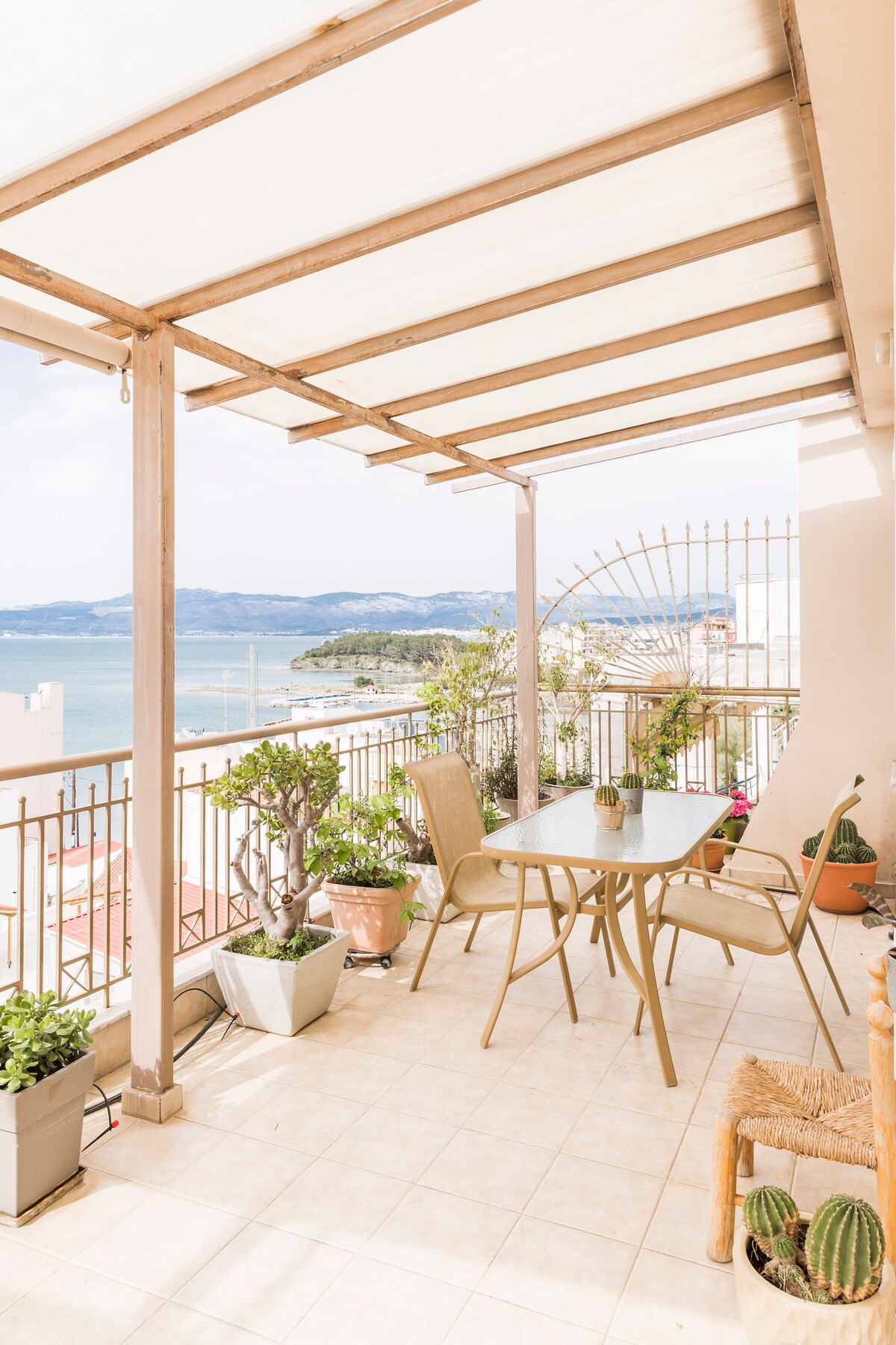 A sunlit balcony features a table and chairs, surrounded by various potted plants. The view includes the coastline and mountains in the distance, framed by a light canopy above. The tiled floor adds a clean and inviting touch to the outdoor space.