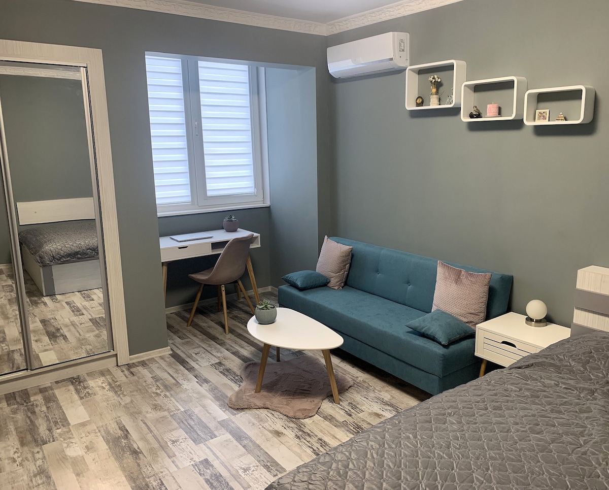 A bright living area features a teal sofa and a low coffee table, with a matching desk positioned beside a window. Natural light filters through the window, illuminating the light gray walls and wooden floor. A mirrored wardrobe adds depth and functionality.