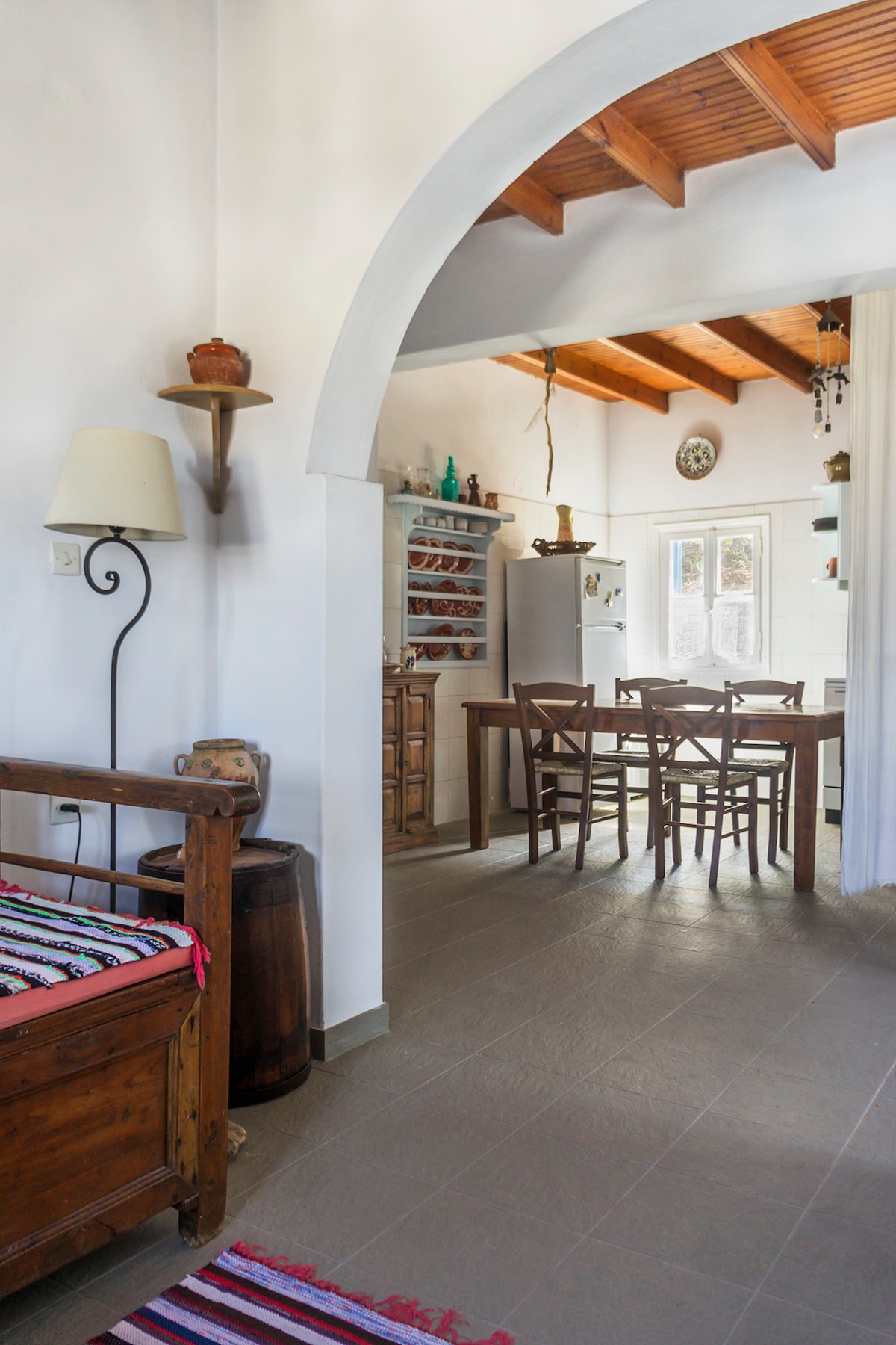 An open living area is defined by high wooden beams and a curved archway. The equipped kitchen is visible, featuring wooden cabinetry and a dining table with six chairs. Decorative items and a warm rug are strategically placed, contributing to the welcoming ambiance.
