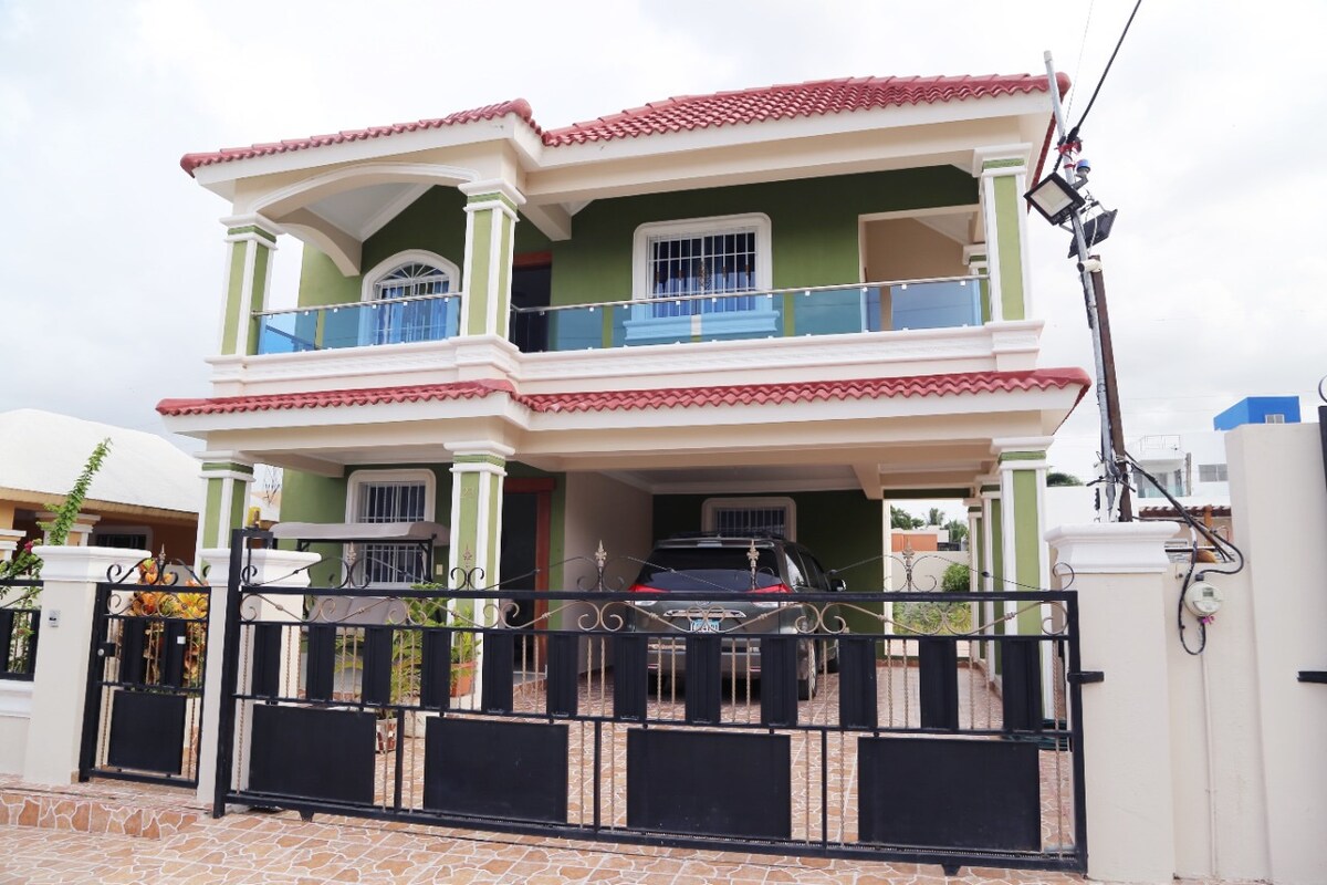 A two-story house with a green exterior features a red-tiled roof and a white balcony railing. A car is parked in the enclosed driveway, which is bordered by decorative stone pavement. Large windows provide natural light, enhancing the inviting facade.