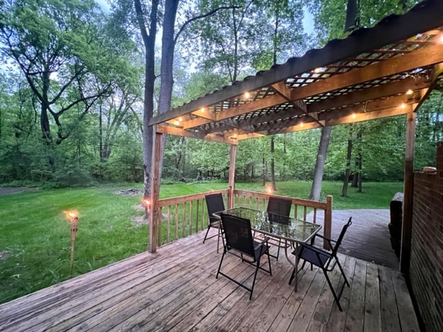 An inviting outdoor deck is visible, featuring a dining table surrounded by black chairs. LED lights are strung overhead, creating a warm atmosphere. The natural surroundings include lush greenery and trees, enhancing the peaceful setting.