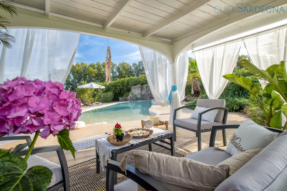 A covered seating area features a comfortable armchair and cushions, with a small table displaying a decorative arrangement. Bright pink hydrangeas and lush greenery frame the view of the tranquil swimming pool and surrounding garden, under soft draped curtains.