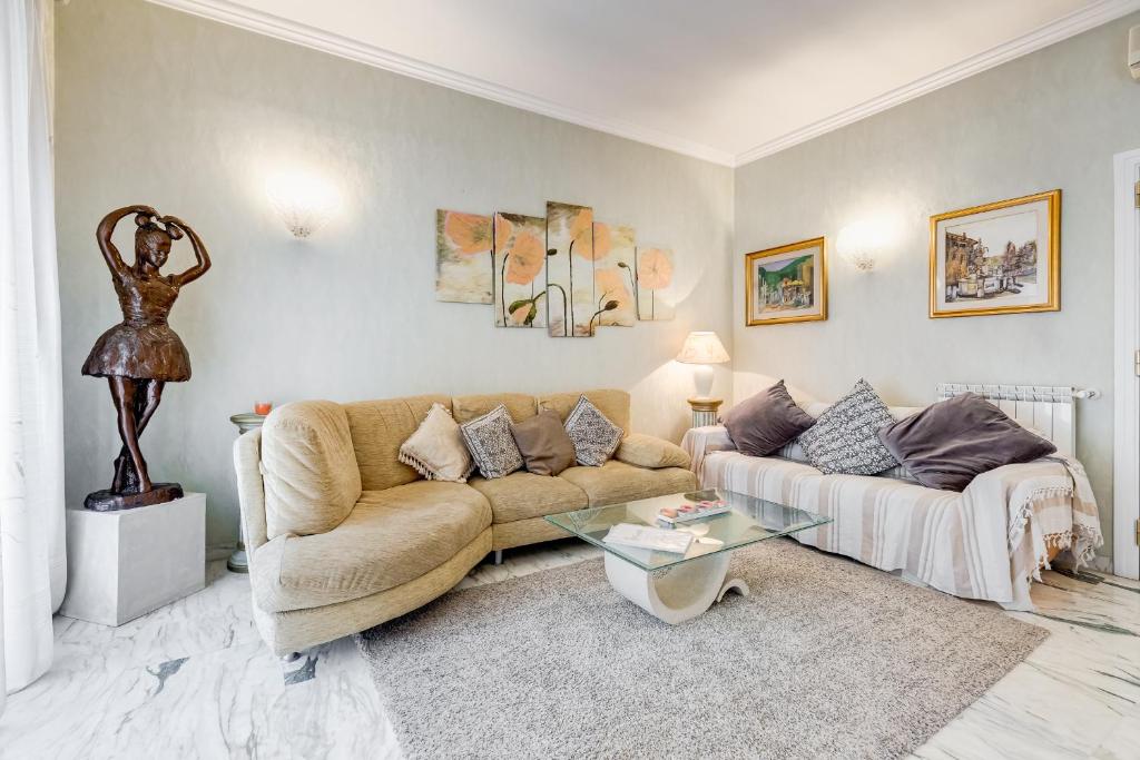 A spacious living area is presented with a curved beige sectional sofa adorned with decorative pillows. A glass coffee table sits on a textured area rug, creating a cozy atmosphere. Art pieces hang on the light green walls, complemented by a sculptural figure and soft lighting.