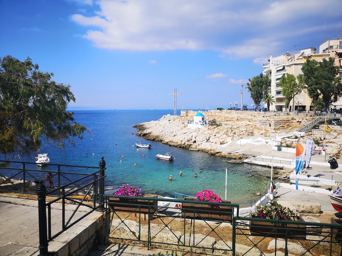 A serene coastal view is presented, featuring clear blue waters lapping against rocky shores. Small boats are anchored nearby, while swimmers can be seen enjoying the water. Vibrant flowers and greenery adorn the waterfront, with buildings visible in the background under a partly cloudy sky.