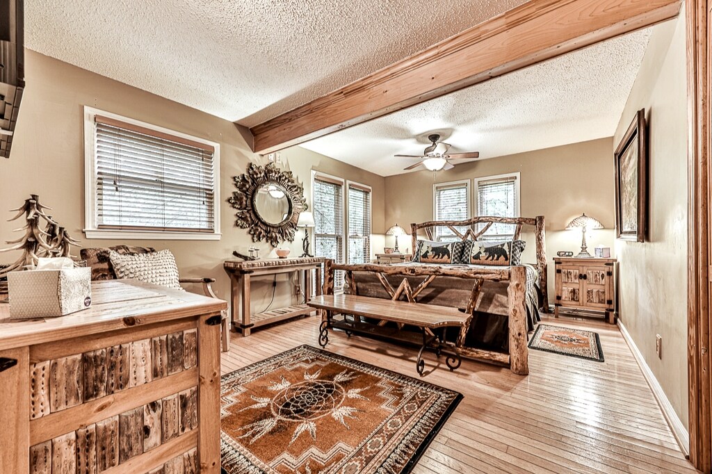 A rustic bedroom features a wooden bed frame and decorative accessories, complemented by warm tones. Natural light filters through multiple windows, illuminating the space. A wooden table and plush area rug enhance the cozy ambiance, with a ceiling fan providing a gentle breeze.