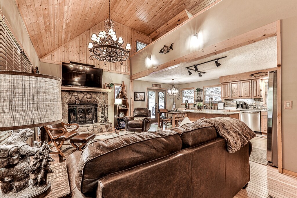 The cozy living area features a leather sofa facing a rustic stone fireplace, with warm wood accents throughout. A chandelier hangs from the high ceiling, and large windows allow natural light to fill the space. The open layout connects to the dining area and kitchen.
