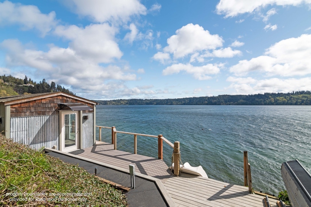 A waterfront deck overlooks calm waters, framed by lush greenery. The wooden structure features large glass doors that open to the outdoor area, providing a seamless view of the landscape. Soft clouds drift across a bright sky, enhancing the serene ambiance.