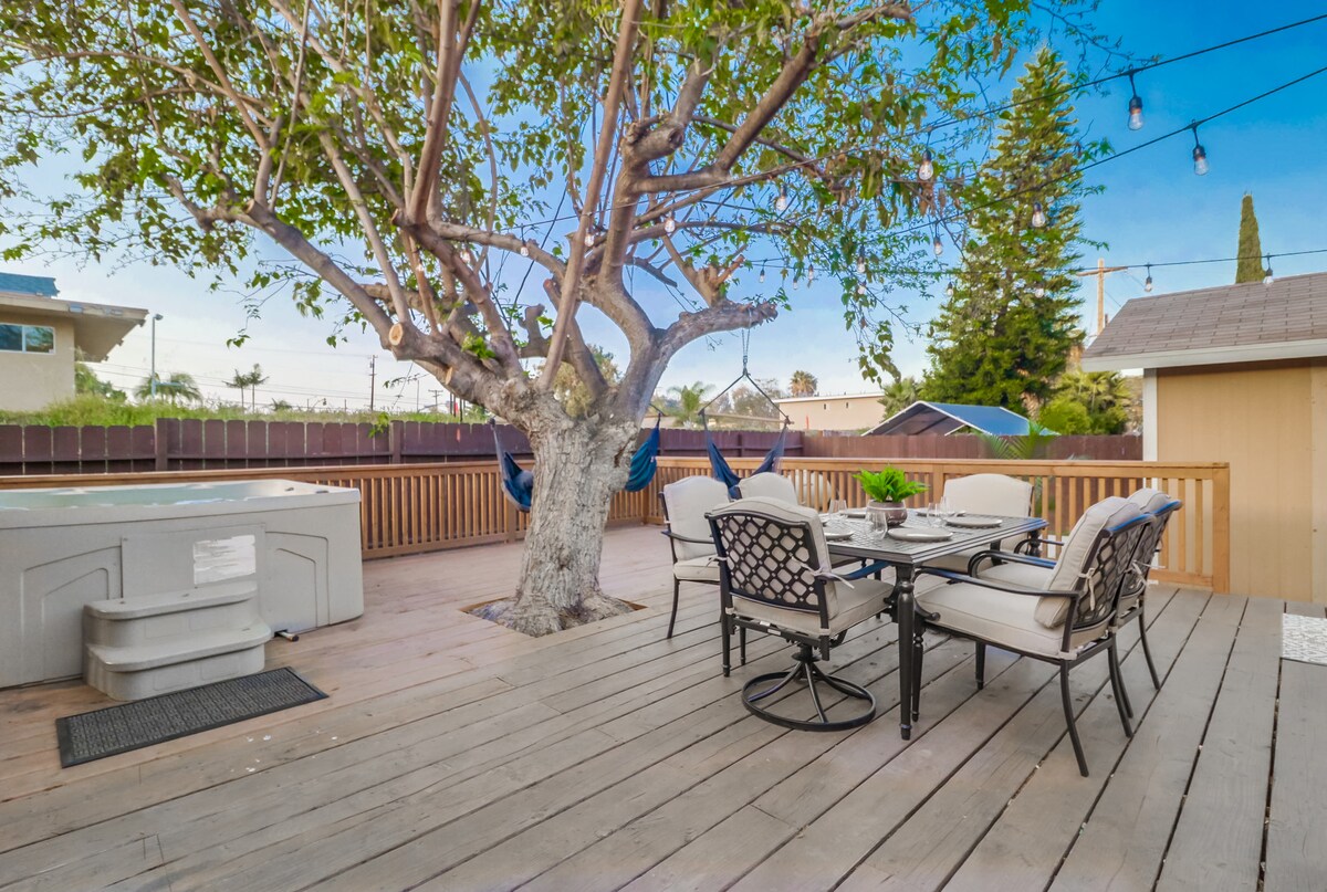An upper deck area features a hot tub beside a dining space, equipped with a table and several chairs. A tree with hanging swings provides shade, and outdoor lighting adds a welcoming touch. Wood decking completes the setting, creating an inviting outdoor environment.
