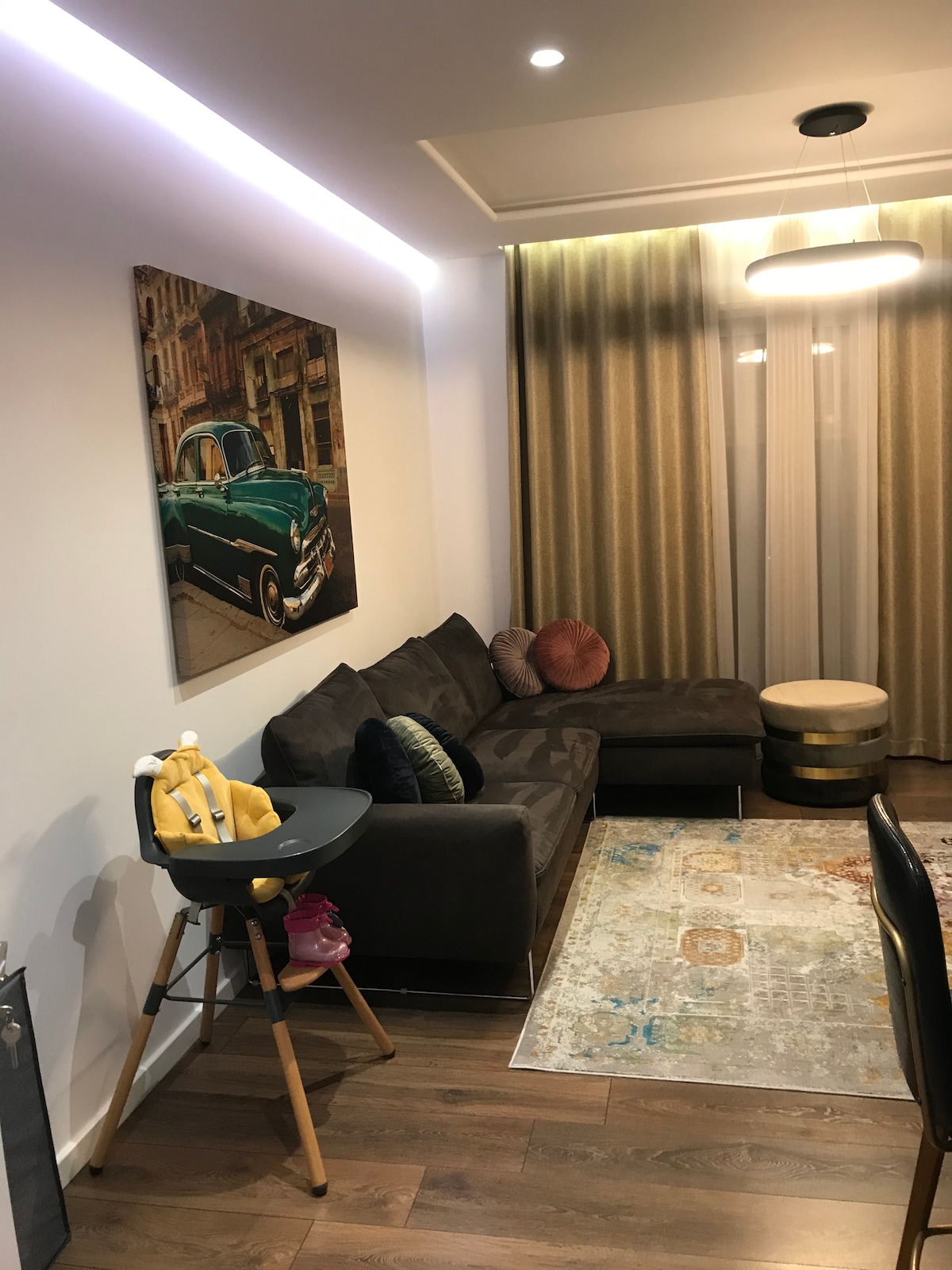 A modern living area features a plush sofa adorned with decorative cushions. A stylish rug lies beneath a circular table, while a vintage car artwork hangs on the wall. Soft, golden drapes frame a large window, allowing natural light to fill the room.