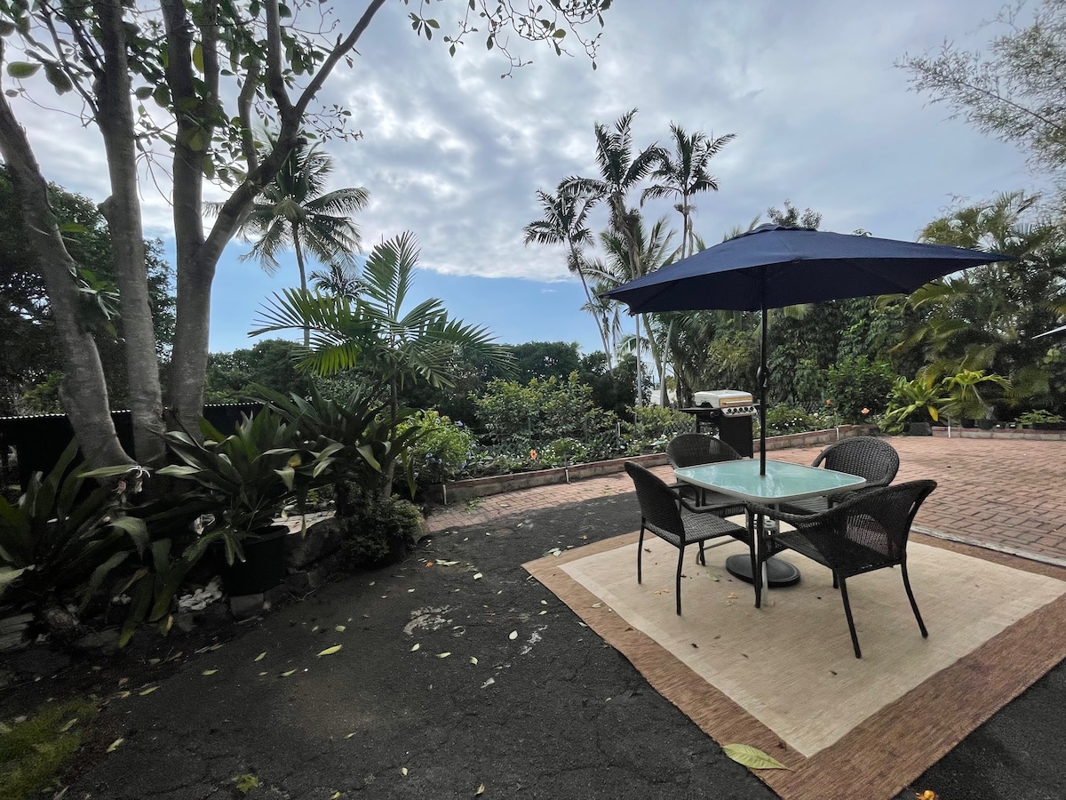 A patio area is shown, featuring a dining table with four chairs under a large blue umbrella. Lush tropical plants surround the space, and a charcoal grill is positioned nearby. A brick pathway leads into the vibrant garden, with a cloudy sky visible above.