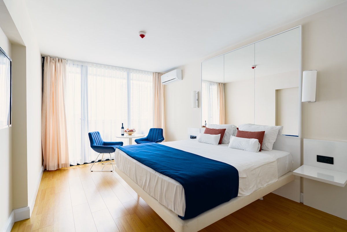 A spacious bedroom is designed with light wooden flooring and neutral-colored walls. A large bed features a white duvet with a blue throw. Stylish chairs and a small table are arranged near floor-to-ceiling windows, allowing natural light to brighten the room.