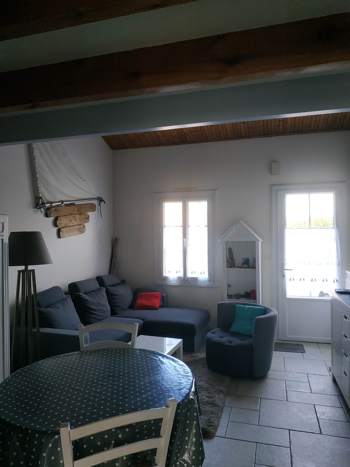 A cozy living area is seen, featuring a comfortable sofa and a small armchair. Light floods through two windows, brightening the space. A round dining table with a polka-dot tablecloth is positioned nearby, while decorative nautical elements adorn the walls, enhancing the room's charm.
