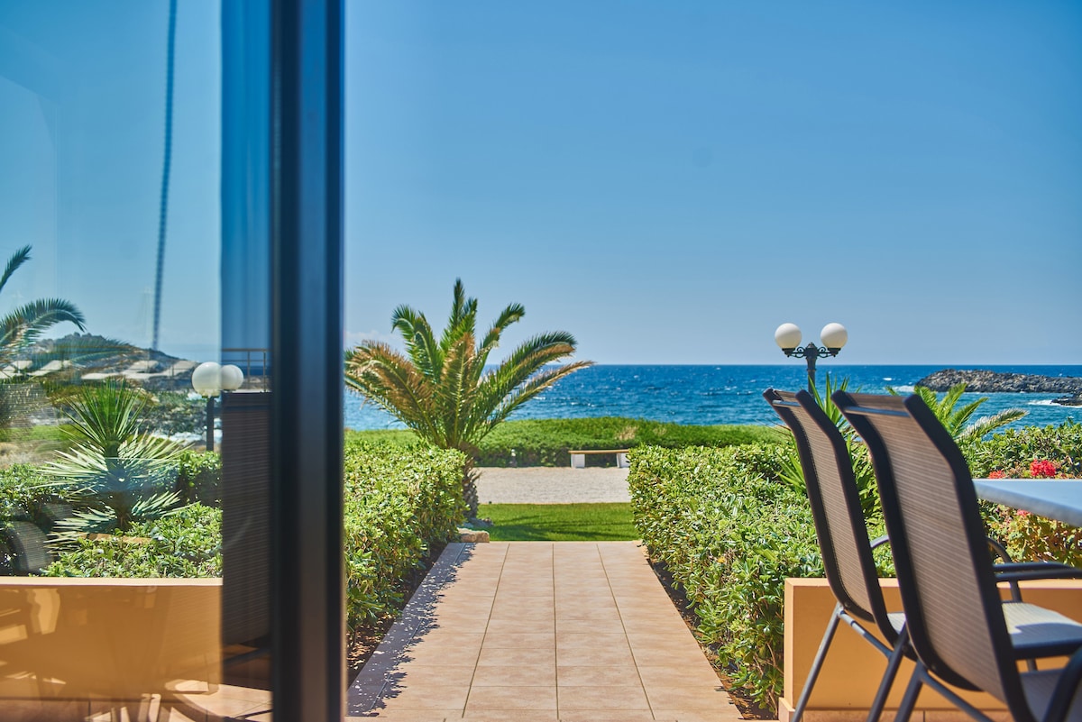 A pathway lined with greenery leads to a view of the sea, framed by palm trees. Outdoor furniture is visible, facing toward the ocean. A bright blue sky and calm waters enhance the tranquil ambience.