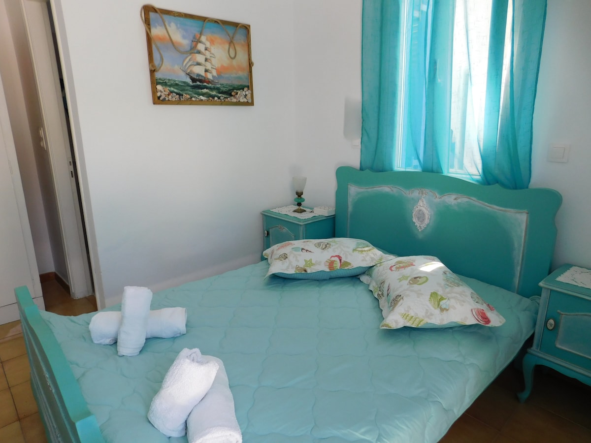 A cozy room is furnished with a double bed adorned with light-colored pillows and soft bedding. Two nightstands in a soothing turquoise hue complement the decor. Natural light enters through a window covered by sheer curtains, while a nautical-themed painting hangs above the bed.