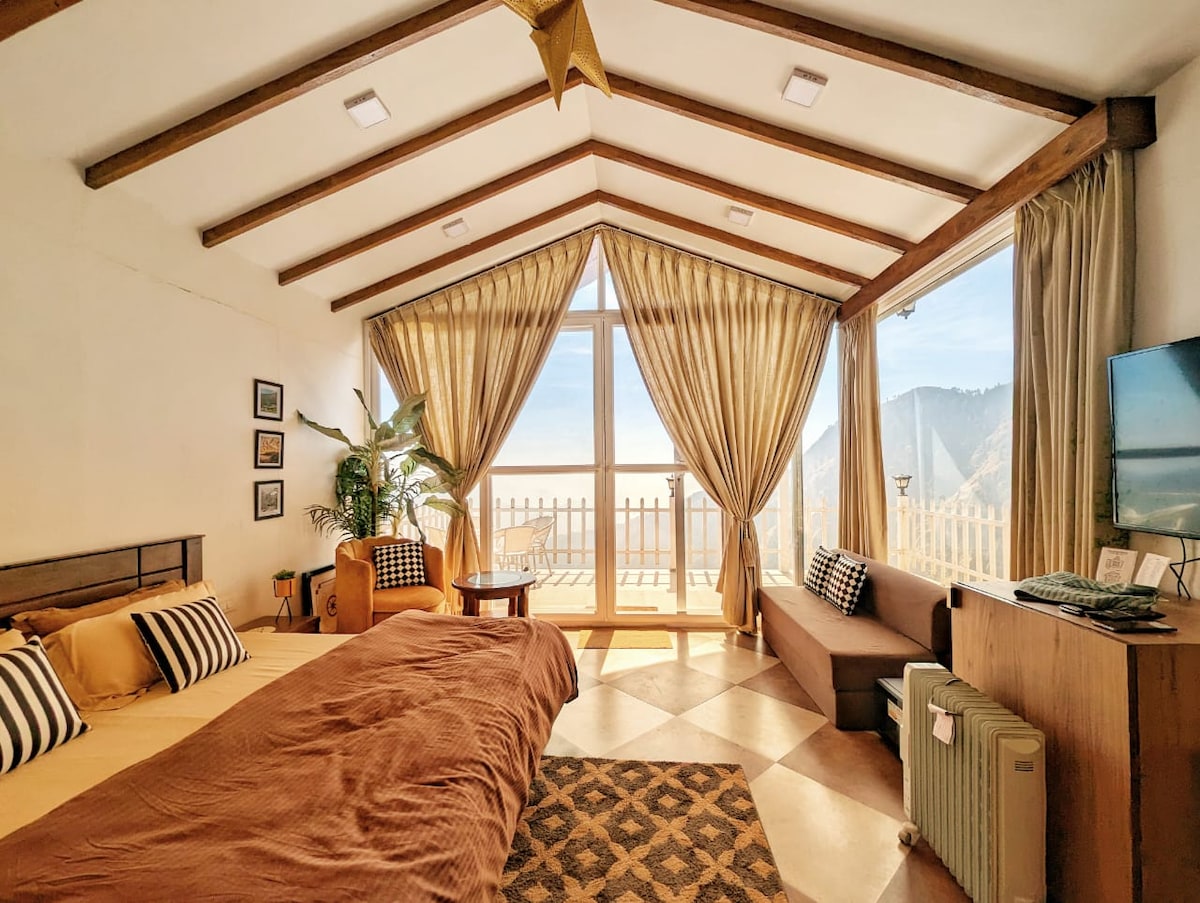 A spacious bedroom features large windows providing views of the surrounding mountains. The room includes a double bed with striped pillows, a sofa, and a decorative rug. A kitchenette, TV, and room heater are visible, while curtains enhance the natural light filtering in.