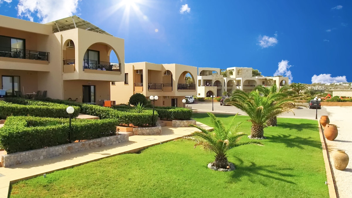 The image displays the Nanakis Beach complex with multiple buildings featuring arched entries. Lush green lawns are accentuated by palm trees and neatly arranged pathways. Ample sunlight highlights the warm tones of the structures against a clear blue sky.