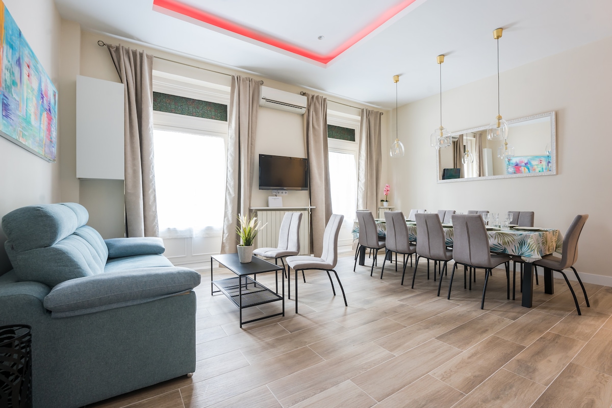 A spacious living and dining area features a comfortable sofa and a dining table suitable for ten. Natural light enters through large windows adorned with light curtains. The ceiling includes modern lighting elements, and a decorative painting enhances the ambiance.