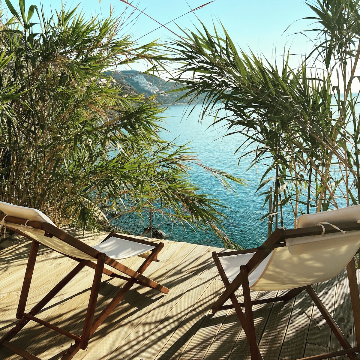 Two wooden lounge chairs are positioned on a wooden deck surrounded by lush greenery, offering a serene view of the calm turquoise sea. The gentle reflection of sunlight on the water creates a tranquil atmosphere, inviting relaxation and peaceful contemplation.