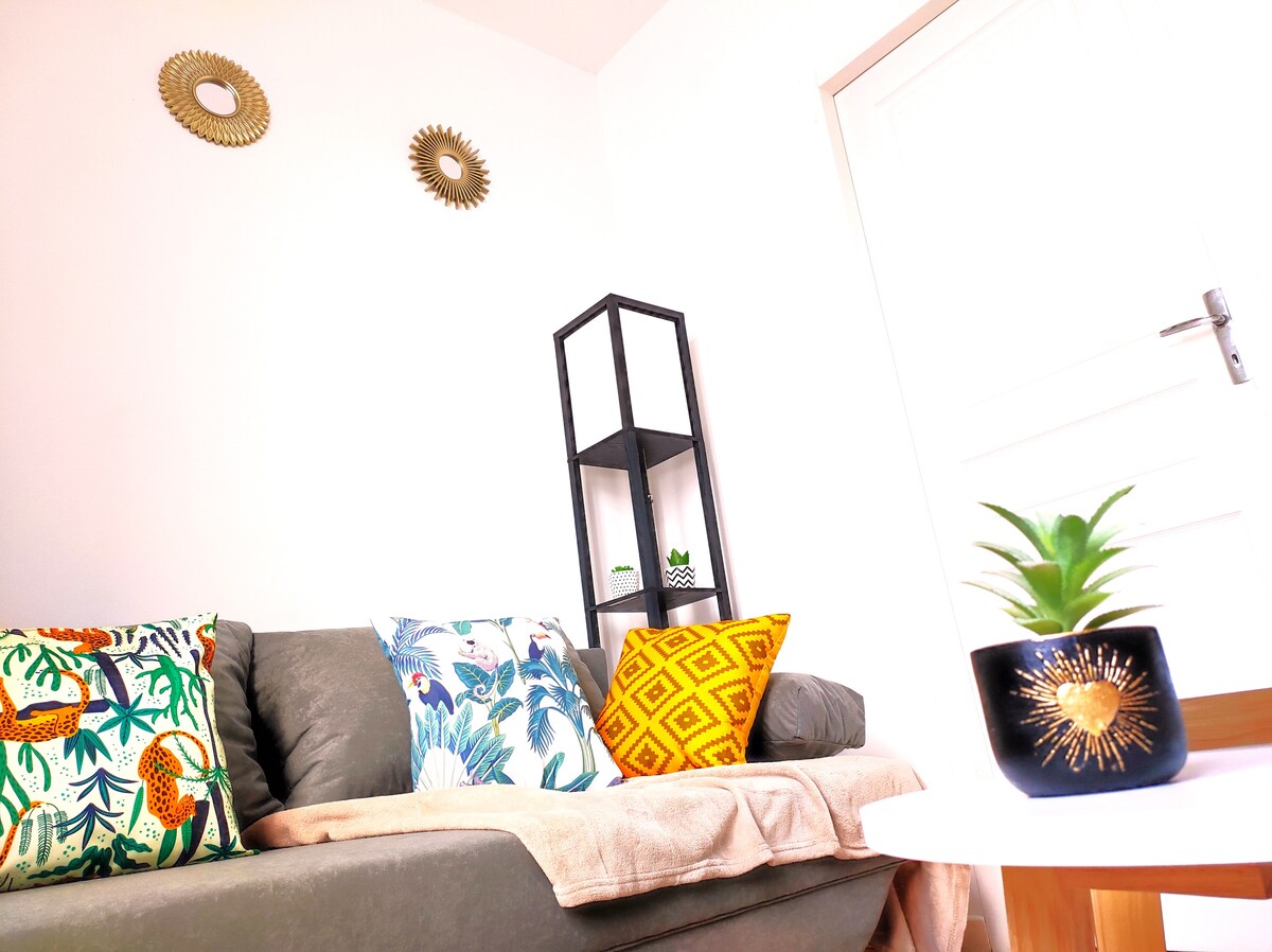 A cozy living area features a comfortable gray sofa adorned with decorative pillows showcasing vibrant patterns. A small plant sits on a round table, while a shelf in the background displays various green plants. Soft light filters through a nearby door.
