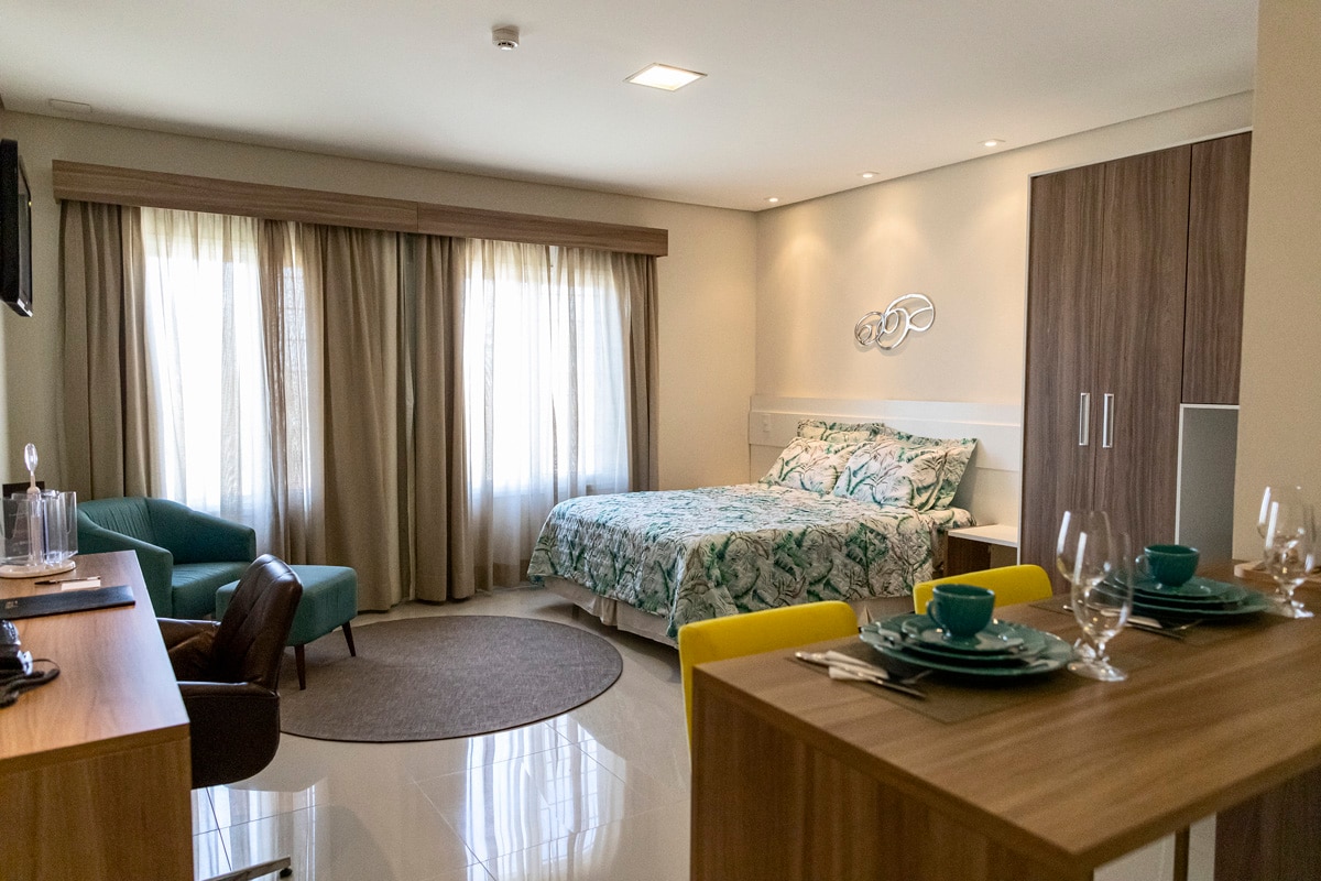 A well-appointed studio features a spacious bed adorned with a floral design. Natural light filters through the large, sheer curtains. A compact dining area and seating options are included, along with a stylish wooden sideboard that complements the modern aesthetic.