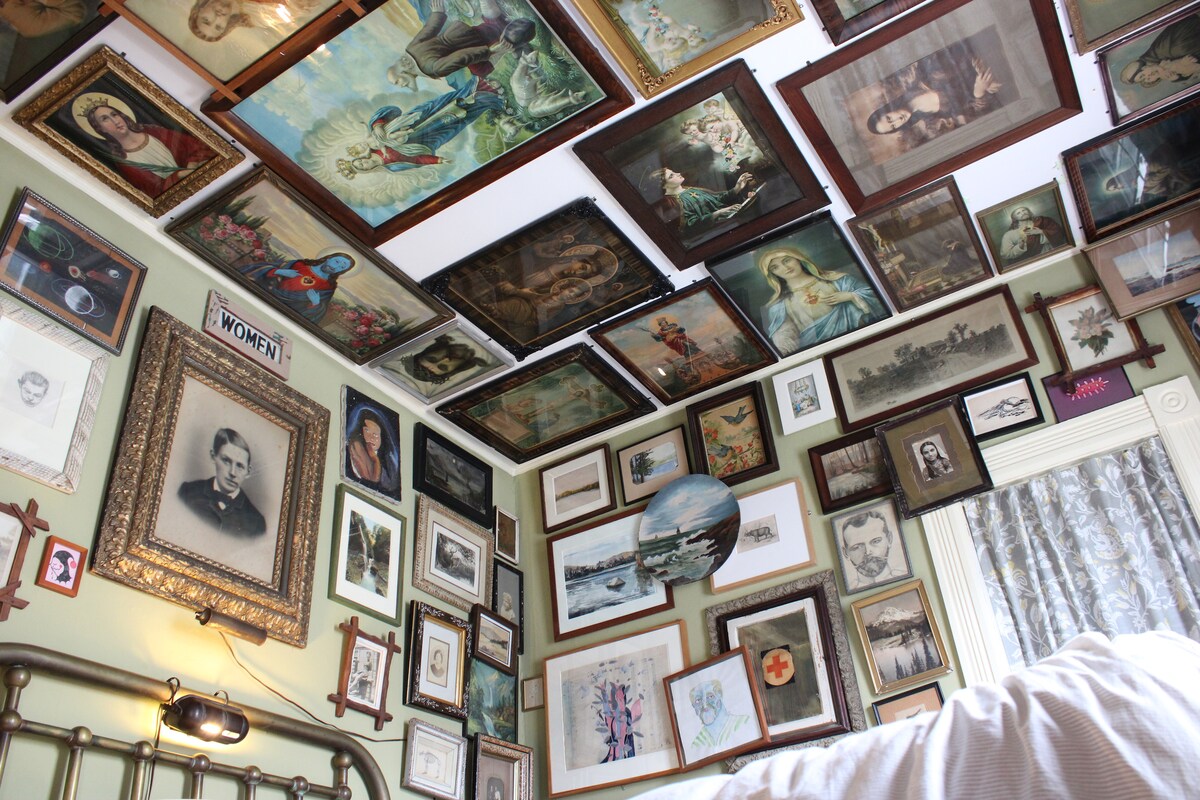 An eclectic collection of framed artwork is displayed on the walls and ceiling of the room. The diverse pieces include portraits, landscapes, and abstract designs, creating a visually engaging environment. A vintage bed frame is visible in the foreground, complementing the artistic atmosphere.