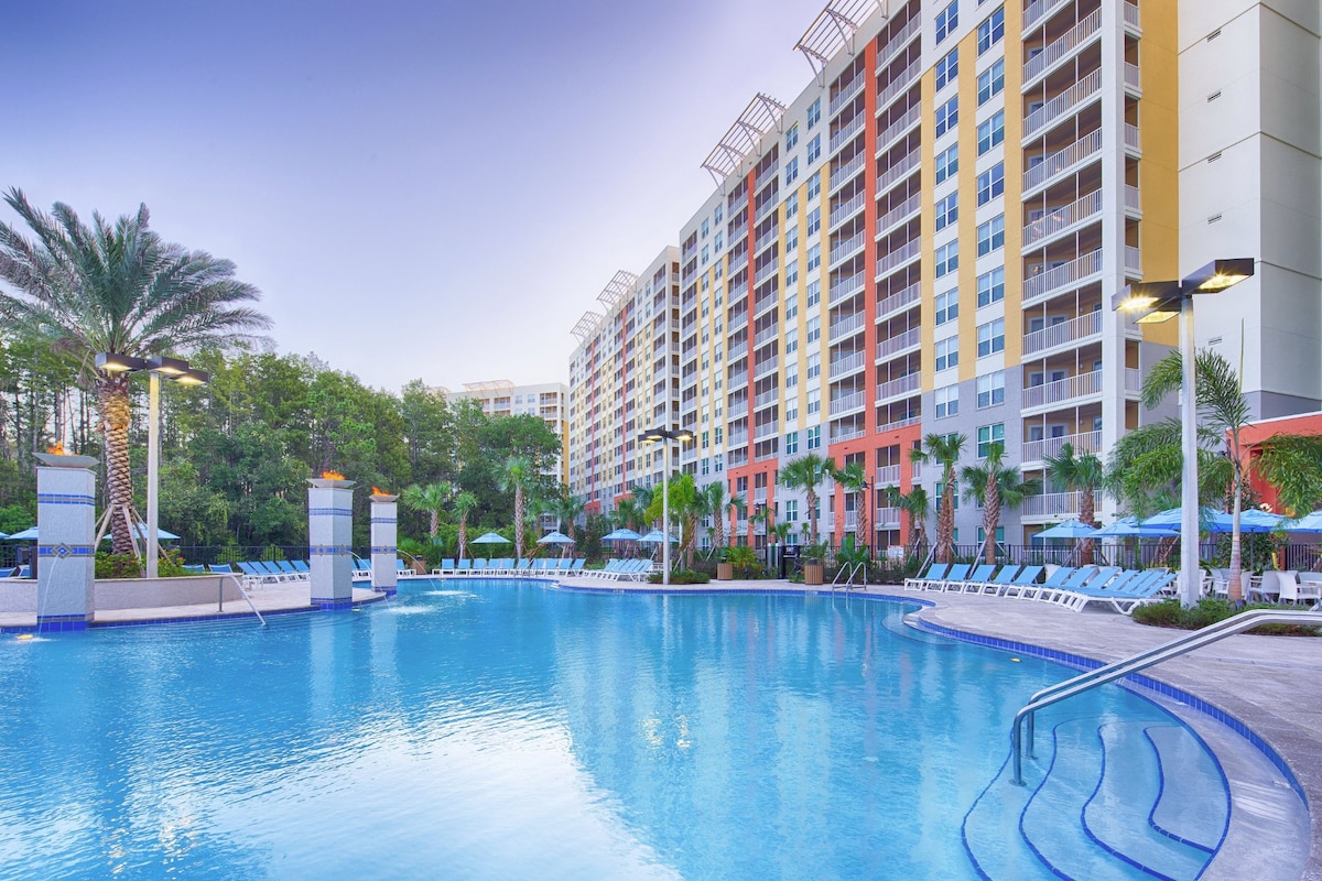 A large outdoor pool is surrounded by palm trees and lounge chairs, providing a spacious relaxation area. Several umbrellas offer shade around the pool, while the multi-story resort building stands in the background, showcasing various colors and architectural details.