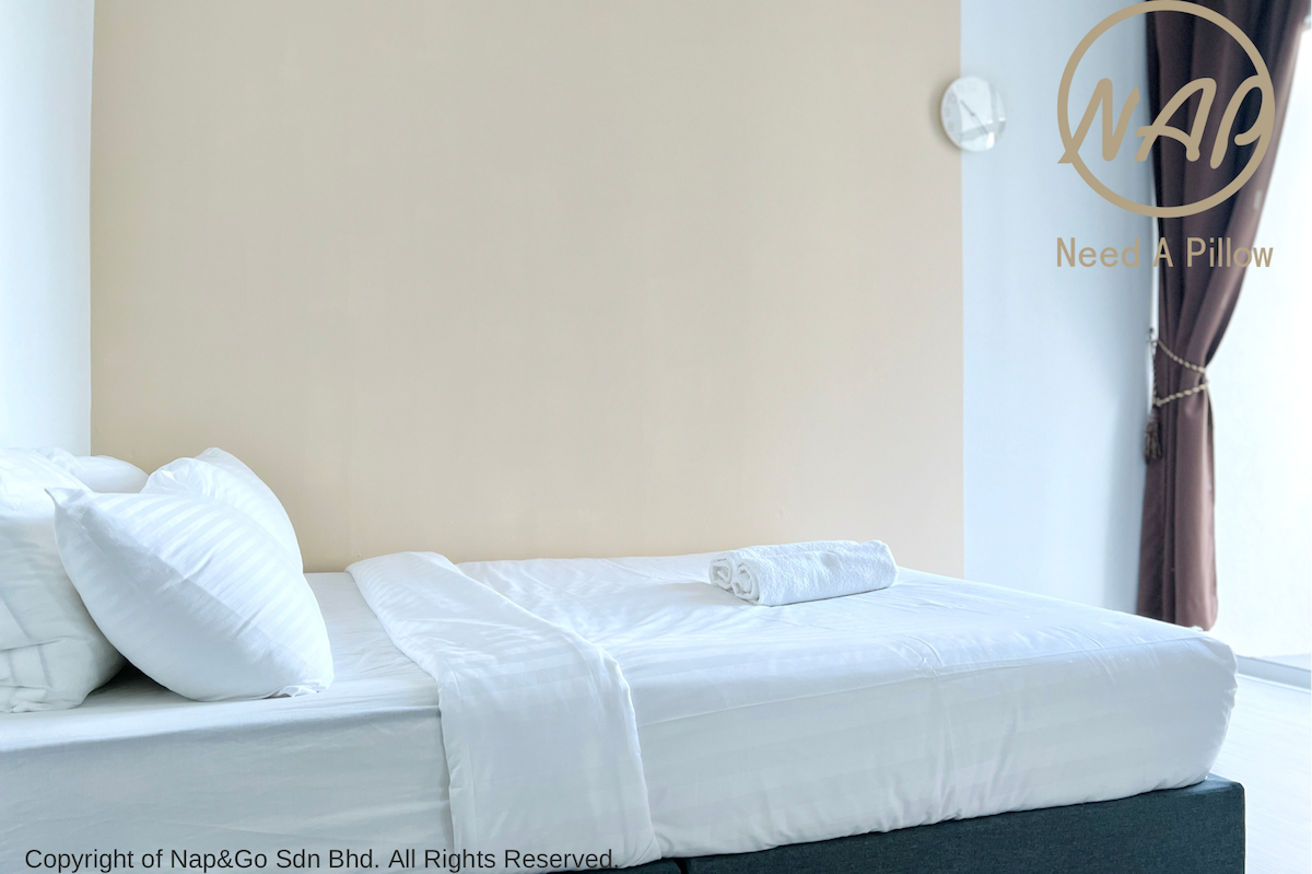 A well-arranged sleeping area features a spacious bed with crisp white linens and a rolled bath towel placed neatly on top. A single pillow rests comfortably, and a wall-mounted clock is positioned nearby. Natural light filters in through a dark-curtained window.