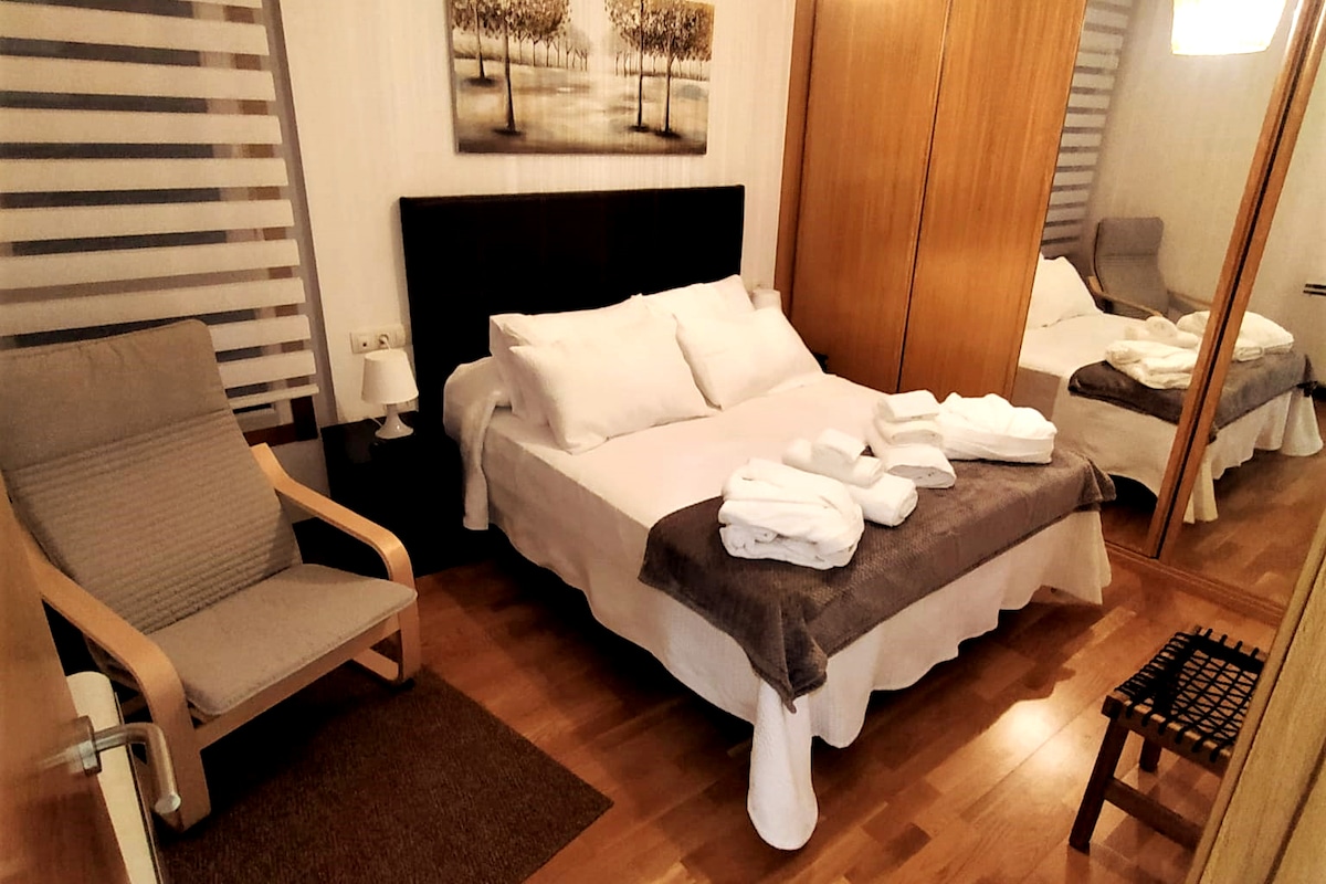 A cozy bedroom features a spacious bed with neatly arranged white linens and towels. A wooden armchair is positioned nearby, and a mirrored wardrobe enhances the sense of space. Soft, natural light filters through striped window coverings.