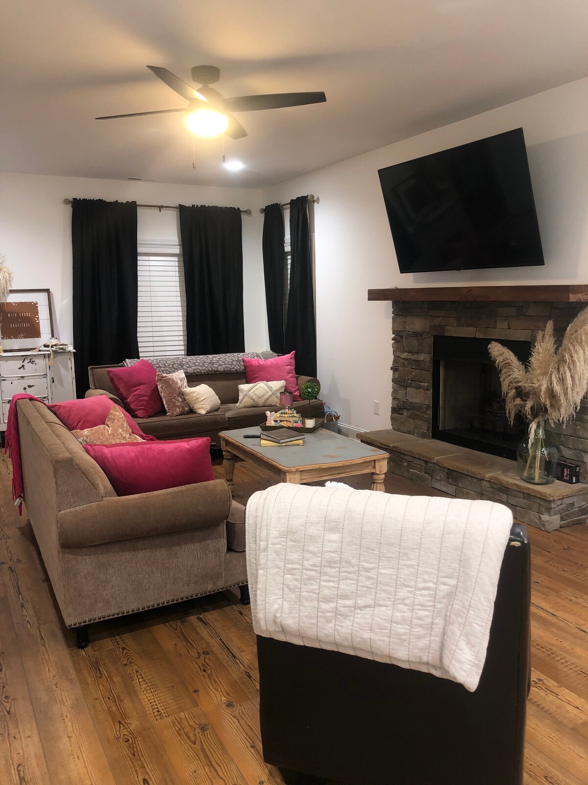 A cozy living area features a sectional sofa adorned with vibrant pink and neutral throw pillows. A coffee table sits in the center, while a stone fireplace provides warmth. Large windows are dressed with dark curtains, and a ceiling fan adds to the comfort.