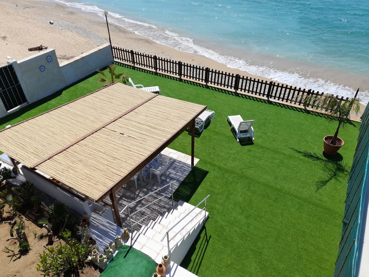 A spacious outdoor area features a covered seating area with a bamboo roof, surrounded by well-maintained artificial grass. Several white lounge chairs are positioned nearby, with a view of the beach and ocean waves gently lapping at the shore.