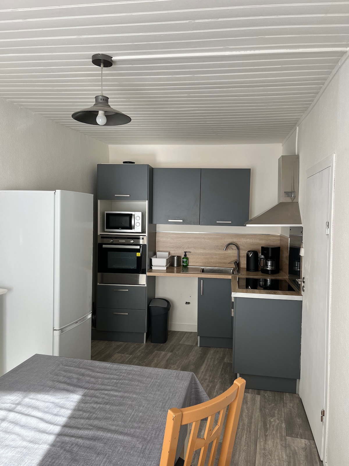 A compact kitchen features modern grey cabinetry and a white refrigerator. Appliances include a microwave and an electric kettle. A wooden dining table with a grey tablecloth is positioned nearby. The ceiling has white paneling, and natural light illuminates the space from a window.