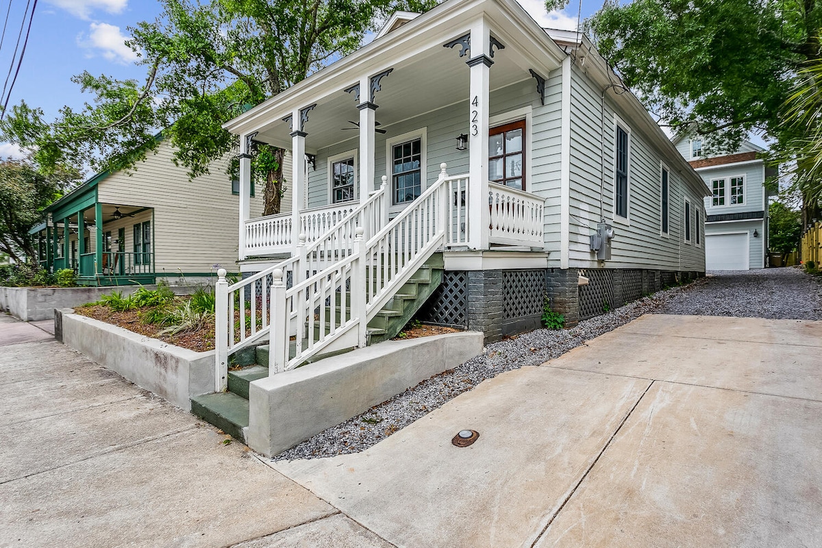 A charming historic home is characterized by its light green exterior and welcoming front porch. White railings and steps lead to the front entrance, surrounded by well-maintained landscaping and stone detailing. Ample driveway space is visible, providing easy access for vehicles.