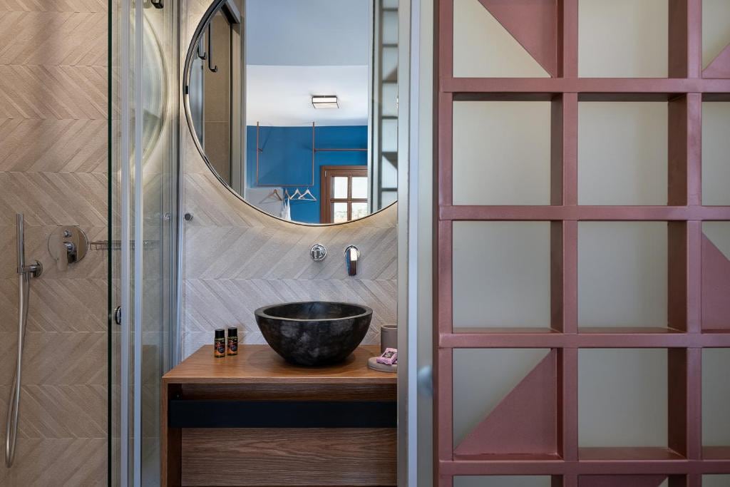 A modern bathroom features a sleek, circular mirror above a dark stone basin, set against a textured wall with herringbone patterns. Dividing the space, a decorative partition showcases a geometric design in muted pink tones, enhancing the room's contemporary feel.