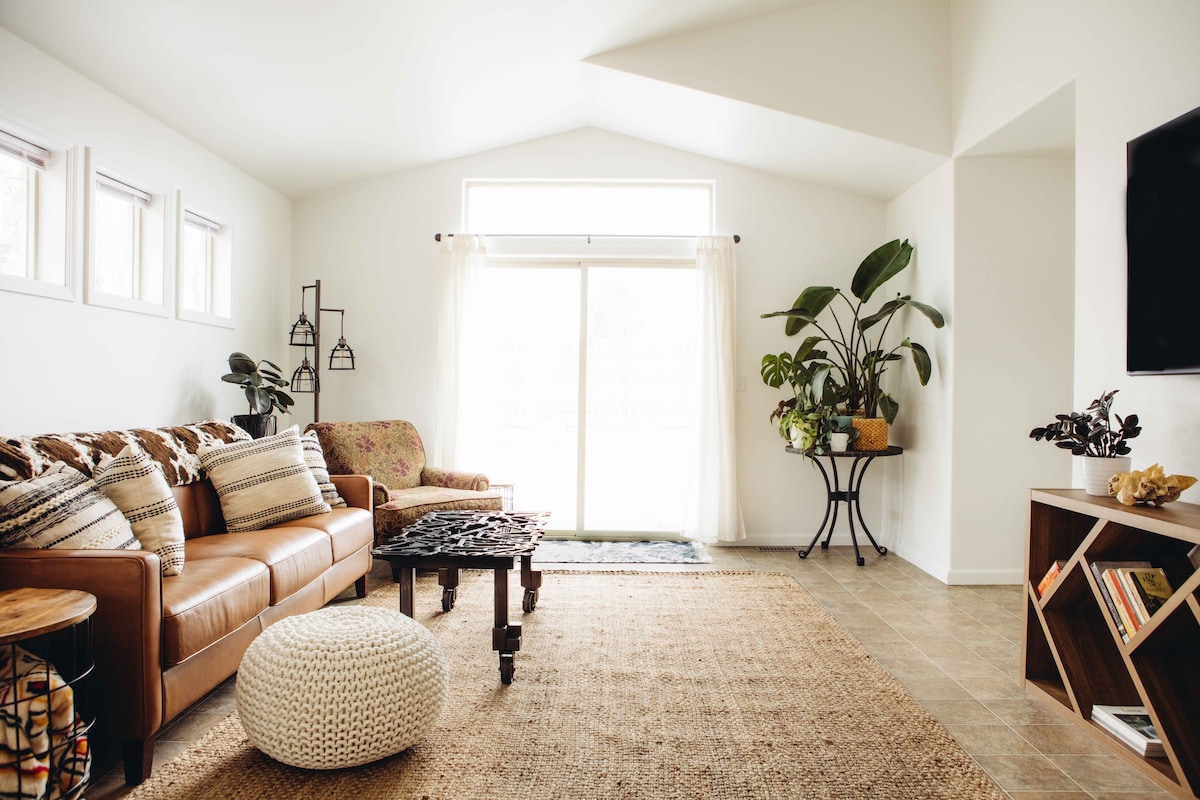 A spacious living area is highlighted by a light-filled window and sliding glass door. A comfortable brown leather sofa and a cozy armchair are positioned near a decorative coffee table. Plants add a touch of greenery, while a textured rug covers the floor.