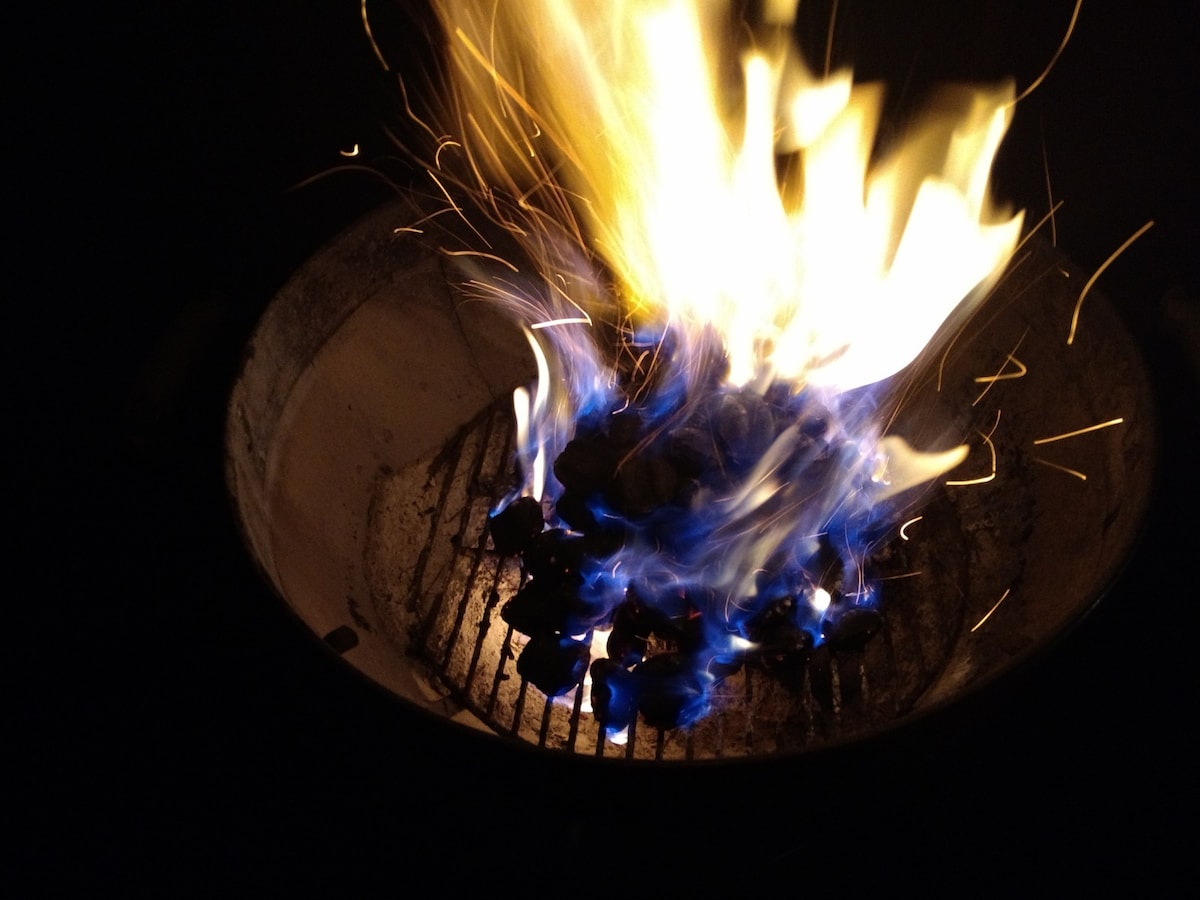 A charcoal grill is shown with vibrant flames rising prominently, surrounded by glowing embers. The dynamic spots of blue and orange in the fire create a striking visual contrast against the dark background, indicating a lively atmosphere for outdoor cooking.