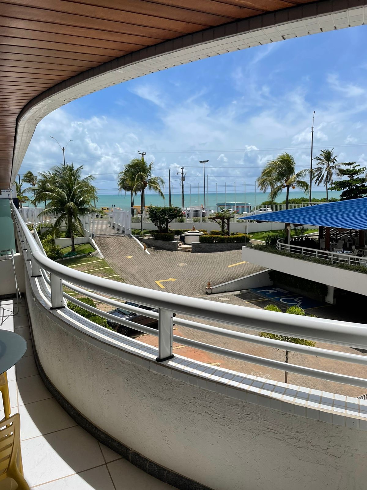 A balcony view showcases the ocean and palm trees, with a clear blue sky overhead. The railing curves elegantly, offering a spacious perspective of the surrounding area, including a nearby parking lot and the shoreline in the distance.