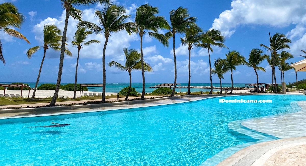 A large swimming pool with clear blue water is featured, surrounded by palm trees swaying in the gentle breeze. The pool area offers panoramic views of the azure sea and a sandy beach in the distance, creating a serene outdoor atmosphere.
