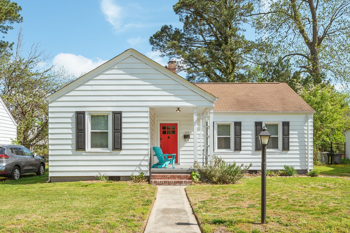 A charming single-story home features a light-colored exterior and a welcoming red front door. A pair of blue chairs is positioned on the porch, with a manicured lawn and shrubs providing a natural setting. A lamp post stands near the walkway leading to the entrance.