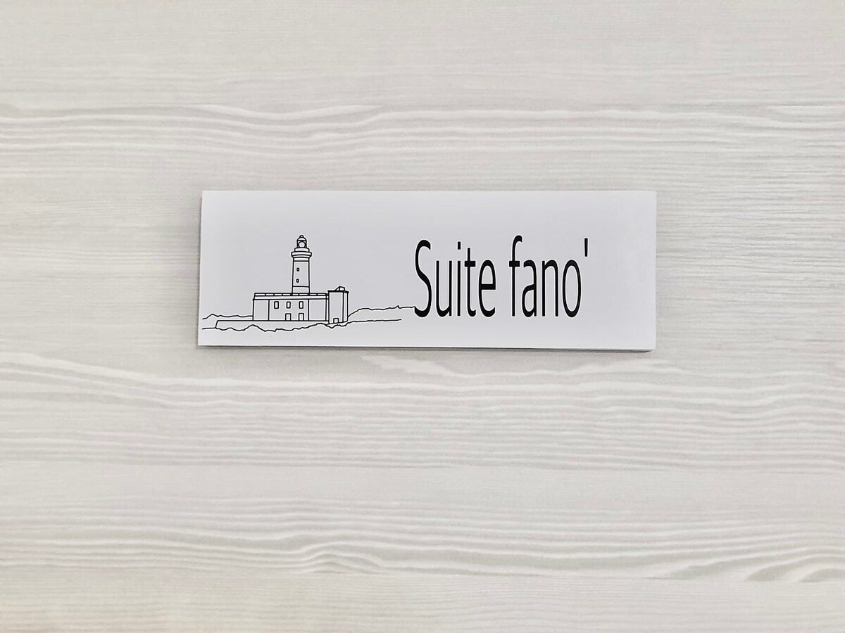 A sign labeled 'Suite Fano' is displayed against a light wooden background, featuring a simple illustration of a lighthouse. The design incorporates a clean, modern aesthetic that emphasizes clarity and elegance.