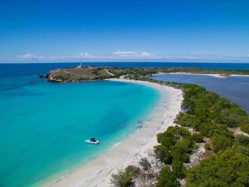 Aerial view captures a scenic stretch of beach with soft, white sand and clear turquoise waters. The landscape features gentle waves lapping at the shore, with a small boat anchored nearby. Lush greenery lines the coastline, while a lighthouse is visible in the distance.